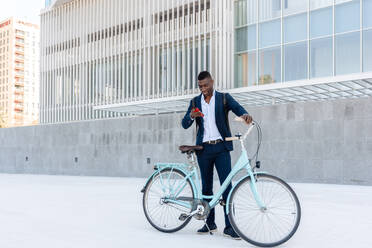 Full body of smiling African American male employee in formal clothes listening to music with headphones while using smartphone and standing near tiled concrete wall looking at screen with bicycle - ADSF44955