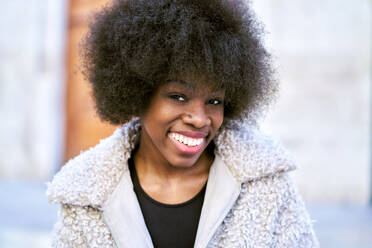 Portrait of cheerful African American young female in cozy clothes with Afro hairstyle smiling and looking at camera while standing against blurred background in daylight - ADSF44958