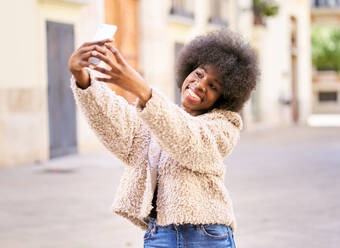 Fröhliche afroamerikanische Frau mit modischer Jacke und voluminösem Haar, die grinst, während sie ein Selfie mit ihrem Handy inmitten einer städtischen Szenerie macht - ADSF44959