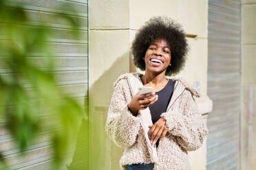 Positive African American female with Afro hairstyle smiling with closed eyes while texting on smartphone and leaning on wall with plant in daylight - ADSF44960