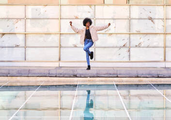 Full body of African American female in casual clothes and Afro hairstyle dancing while jumping and having fun on terraced floor beside bright framed wall and reflective pool of water in daylight - ADSF44965