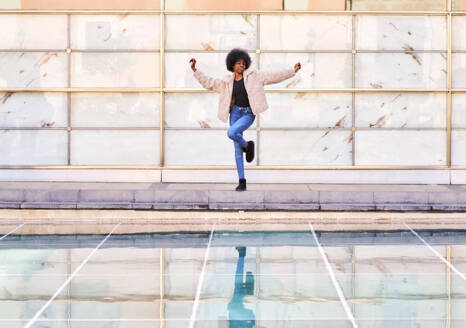 Full body of African American female in casual clothes and Afro hairstyle dancing while jumping and having fun on terraced floor beside bright framed wall and reflective pool of water in daylight - ADSF44965