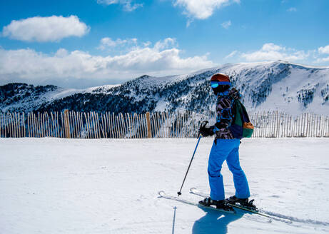 Full body side view of unrecognizable skier in warm ski suit and with skiing equipment standing on snowy slope with skiing poles - ADSF45000