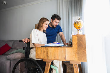 Adorable loving husband assisting wife sitting in wheelchair with office project at home using laptop - ADSF45010