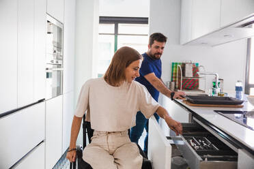 Adorable husband with wife sitting in wheelchair in casual clothes happily discussing while preparing meal together in modern kitchen - ADSF45013