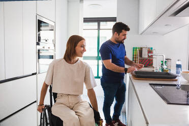 Caring husband in casual clothes helping happy disabled wife with preparing food together in kitchen while having fun - ADSF45014