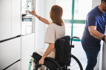 Side view of unrecognizable husband in casual clothes with disabled wife in wheelchair using modern equipment in hospital - ADSF45015