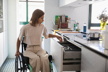 Busy young female in casual clothes sitting on wheelchair by drawer while doing house chores in kitchen in modern apartment - ADSF45017