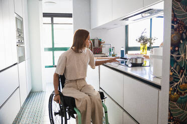 Busy young female in casual clothes sitting on wheelchair by drawer while doing house chores in kitchen in modern apartment - ADSF45018