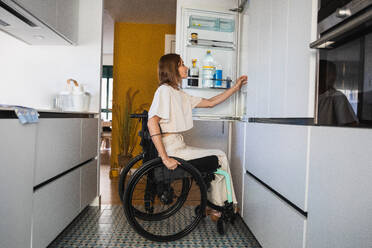 Side view of young disabled female in casual clothes sitting on wheelchair with hand on opened fridge in kitchen at daylight - ADSF45019