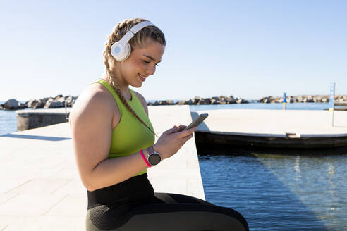 Side view of fit female athlete in sportswear listening to music in wireless headphones and browsing mobile phone while sitting on embankment near sea - ADSF45028