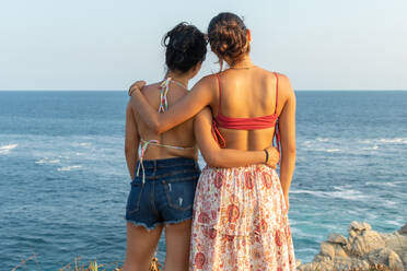 Back view of anonymous female friends in swimwear and summer clothes embracing while standing on stony coast against waving sea during vacation together in Puerto Escondido - ADSF45029