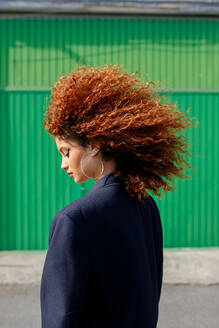 Side view of young female model with curly hairstyle and closed eyes wearing black jacket and standing against green wall - ADSF45033