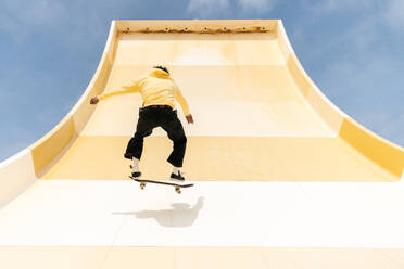 Back view of full body African American male skater in casual clothes holding skate board while running on ramp in skate park at countryside in daylight during training - ADSF45042