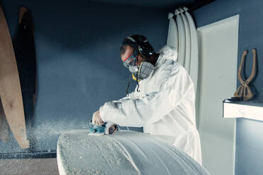 Side view of focused male master in protective respirator and uniform using professional equipment for sanding surfboard surface while working in modern workshop - ADSF45056
