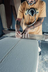 Anonymous tattooed male master in yellow t shirt with respiratory mask on neck using special measuring tool and pencil for making marks on surfboard during work process in professional workshop - ADSF45057