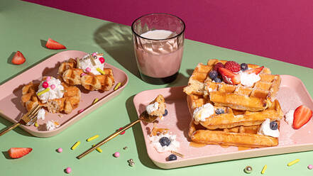 From above tasty sweet waffles topped with berries fruits sauce and cream served on pink tray on colorful table background near glass with hot milk beverage in light kitchen - ADSF45058