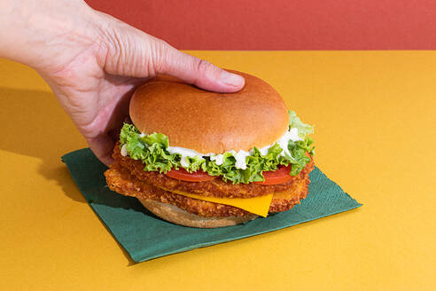 Cropped unrecognizable person eating delicious fish burger with tomatoes, cheese slices and fresh lettuce on yellow and red background - ADSF45068