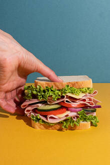 Cropped unrecognizable person holding delicious sandwich with ham, tomatoes, cucumber and cheese slices and fresh lettuce on blue and yellow background - ADSF45070