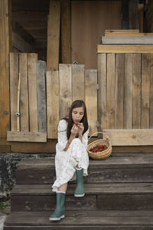 Girl examining strawberry sitting by basket on steps - LESF00344