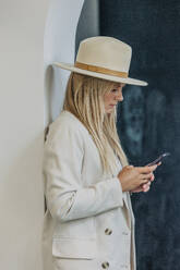 Blonde Frau mit Smartphone in der Nähe der Wand stehend - VSNF01150