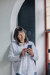 Senior woman using smart phone leaning on wall - VSNF01159