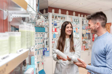 Positive young female pharmacist in white coat with cheerful man each holding bottle of chemical liquid while discussing in pharmaceutical shop with drugs on shelves - ADSF45073
