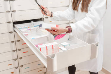 Crop side view of young female scientist in white laboratory coat standing by slotted shelves with drugs while using tablet in modern pharmacy store - ADSF45076