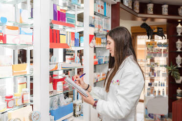 Side view young female pharmacist standing and searching for medicines on shelves using notebook in drugstore - ADSF45078