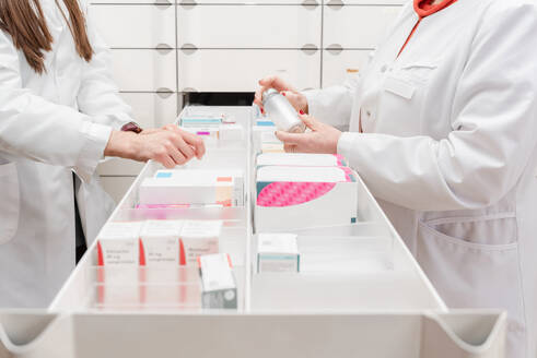 Side view of female pharmacist interacting with female colleague while organizing medicines in cabinet - ADSF45079