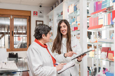 Happy young female pharmacists in white medical uniform smiling and taking notes on notebook while browsing tablet and standing near shelves with medical supplies in pharmacy - ADSF45082