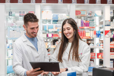 Young pharmacists in white robe standing near shelves with various medical supplies and looking at tablet screen while working in pharmacy - ADSF45086