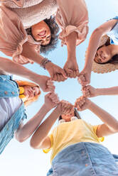 From below group of happy multiracial female friends smiling and looking at camera while standing and stacking hands in daylight during holiday - ADSF45092