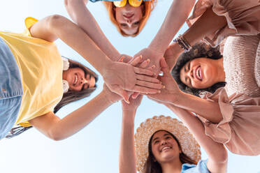 From below of group of happy young multiracial females smiling and looking at camera while standing and stacking hands together in daylight of summertime - ADSF45094