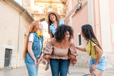 Group of happy multiracial females smiling with one friend having piggyback ride looking at each other while standing on Valencia city street - ADSF45098