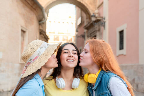 Happy girlfriends smiling and kissing looking up brunette female while standing on Valencia city street with blurred buildings - ADSF45101