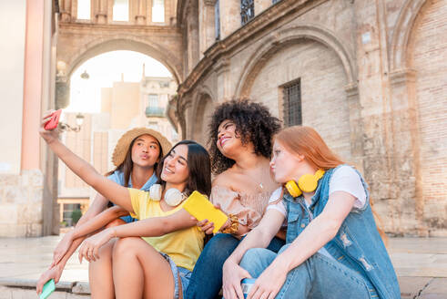 Happy young multiracial girlfriends in casual clothes smiling and looking at mobile screen while sitting on stone and shooting self portrait together against old building in Valencia - ADSF45105