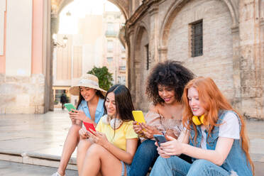 Happy multiracial female friends in stylish clothes sitting on border and browsing on mobile phone while enjoying time together in Valencia - ADSF45108