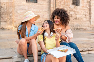 Happy multiracial girlfriends sitting on step smiling and looking at each other while having lunch with pizza together against building in Valencia - ADSF45110