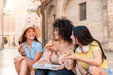 Happy multiracial girlfriends sitting on step smiling and looking at each other while having lunch with pizza together against building in Valencia - ADSF45111