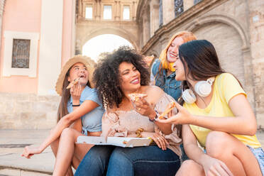 Happy multiracial girlfriends sitting on step smiling and looking at each other while having lunch with pizza together against building in Valencia - ADSF45112