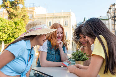 Happy multiracial girlfriends smiling and surfing mobile phone while sitting at table in outdoor cafe during daytime in Valencia - ADSF45113