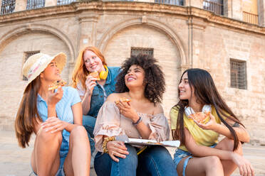 Happy multiracial girlfriends sitting on step smiling and looking at each other while having lunch with pizza together against building in Valencia - ADSF45114