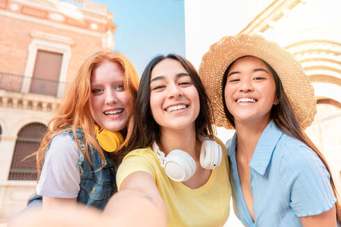 Happy multiracial young girlfriends in stylish outfits shooting self portrait while standing against buildings and looking at camera in Valencia - ADSF45121