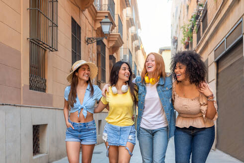 Cheerful multiethnic female friends hugging each other with toothy smile while walking along street with buildings in Valencia - ADSF45127