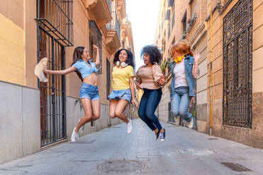 Full body of young happy multiethnic girlfriends laughing and jumping together while having fun during walk in street of Valencia - ADSF45128