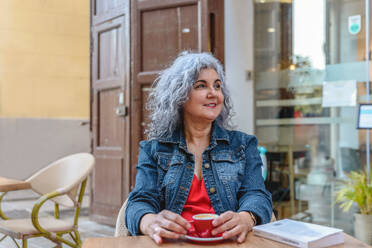 Cheerful elderly female in denim jacket with curly gray hair sitting at table with cup of hot aromatic coffee and looking away while resting in outdoor cafe - ADSF45143