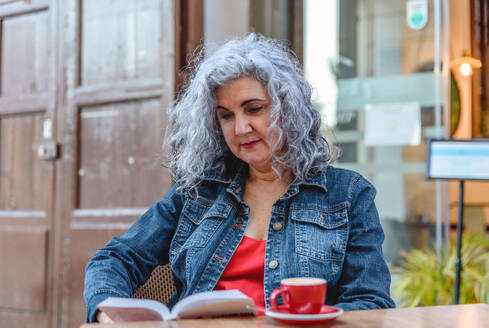 Concentrated senior female in denim jacket and red dress with curly gray hair sitting at table with cup of aromatic coffee and reading book in outdoor cafe - ADSF45144