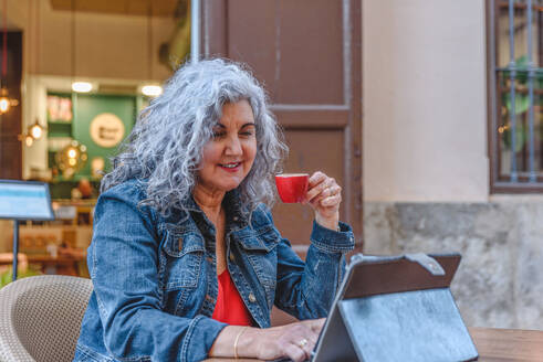 Positive elderly female in casual outfit with curly gray hair smiling and using tablet while drinking hot coffee in outdoor cafe - ADSF45146