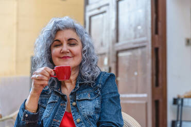 Positive elderly female with eyes closed in casual outfit with curly gray hair smiling while smelling hot coffee in outdoor cafe - ADSF45147